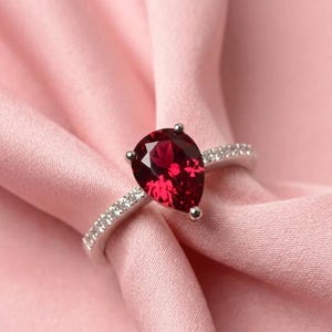 Op de afbeelding: Een zilverkleurige ring met een grote, peervormige, dieprode edelsteen als middelpunt. De ringband is slank en versierd met een rij kleine, heldere, fonkelende steentjes die aan beide zijden van de band zijn ingelegd tot aan de hoofdzetting.