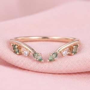 May include: A rose gold ring with a curved design. The ring features green marquise-cut gemstones and small, round, clear gemstones. The ring is set on a pink fabric background.