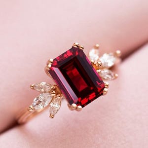 Puede incluir: Un anillo de oro rosa con una gran piedra preciosa granate de color rojo intenso, corte esmeralda rectangular en el centro, flanqueada por tres pequeños diamantes blancos claros de corte marquesa a cada lado, montados en una banda fina pulida.