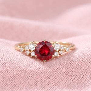 Könnte beinhalten: Ein goldener Ring mit einem großen, runden, tiefroten Edelstein als Mittelpunkt. Der Ring ist mit Clustern kleinerer, klarer Edelsteine auf beiden Seiten des zentralen Steins versehen, vor einem weichen rosa Hintergrund.