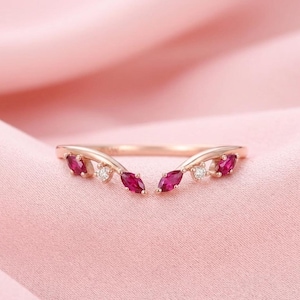 May include: A rose gold ring with a curved design, featuring marquise-cut ruby gemstones and small round diamonds. The ring is set against a soft pink background, showcasing its elegant and delicate details.
