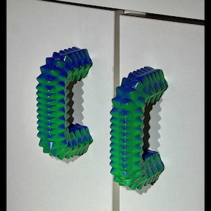 Peut inclure: Deux poignées de placard en forme de C avec un design texturé et pointu. Les poignées sont un dégradé de bleu et de vert, montées sur une porte d'armoire blanche. La quincaillerie est un élément de design moderne.