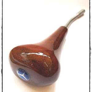 Peut inclure: Un pommeau de levier de vitesse en bois de ronce brun foncé poli avec une finition brillante et une tige en métal argenté. Le dessus du pommeau présente un emblème circulaire bleu avec un logo en étoile argentée à trois branches.