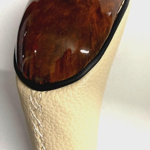 Puede incluir: Un pomo de palanca de cambios con una parte superior de madera pulida en tono marrón y un emblema circular de BMW. La sección inferior está cubierta de cuero beige texturizado con costuras cruzadas blancas, separada por un borde negro.