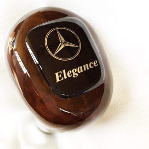 Può includere: Pomello del cambio in legno per auto con finitura lucida. Il pomello presenta un motivo a venature del legno marrone scuro. Un inserto rettangolare nero presenta il logo Mercedes-Benz e la parola "Elegance" in lettere dorate.