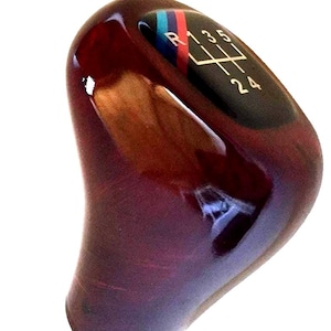 May include: A wooden gear shift knob with a glossy, dark reddish-brown finish. The top features a black panel with white gear shift pattern markings and a multi-coloured stripe. The knob is designed for a vehicle.