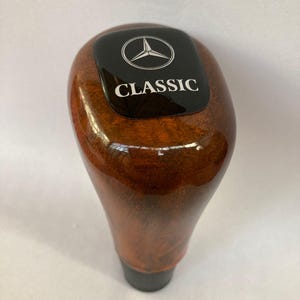Może przedstawiać: Drewniana gałka zmiany biegów z błyszczącym wykończeniem. Na górze znajduje się czarny prostokątny emblemat z logo Mercedes-Benz i białym napisem "CLASSIC". Gałka ma ciemnobrązowy kolor.