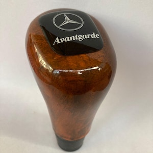 Puede incluir: Pomo de palanca de cambios de madera con acabado brillante. La parte superior del pomo tiene una placa negra con el logotipo de Mercedes-Benz y la palabra "Avantgarde" en blanco. El pomo es marrón con un patrón de vetas de madera.