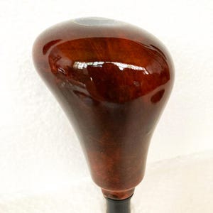 Op de afbeelding: Een glanzende houten versnellingspookknop met een rijke, donkerbruine afwerking. De knop heeft een afgeronde bovenkant en een taps toelopende basis met een zwarte rubberen huls. De houtnerf is zichtbaar en voegt textuur toe aan het gladde oppervlak.