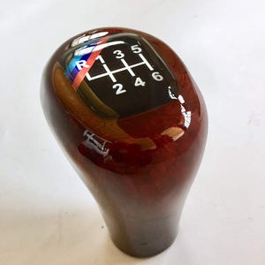 May include: A glossy, dark wood-grain shift knob with a black top featuring a gear pattern. The gear pattern includes numbers 1 through 6, plus reverse. The knob has a smooth, ergonomic shape.