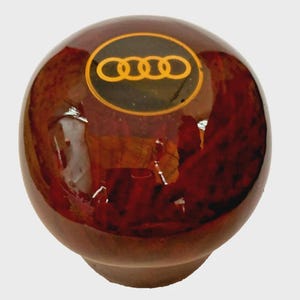 Può includere: Un pomello del cambio rotondo in legno con finitura lucida. Il pomello è di un ricco colore marrone con una venatura scura e modellata. La parte superiore presenta il logo Audi in oro su sfondo nero. Il pomello ha una base arrotondata.