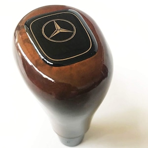 Può includere: Pomello del cambio in legno con finitura lucida. La parte superiore presenta un quadrato nero con il logo Mercedes-Benz in oro. Il pomello ha un motivo a venature del legno marrone e beige e una base argentata.