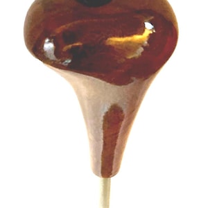 May include: A polished brown wooden gear shift knob with a smooth, rounded top featuring a circular blue and silver Mercedes-Benz logo emblem. The knob tapers downwards into a narrow base connected to a thin, light-coloured metal rod extension.