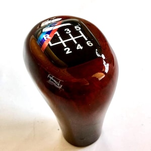 Può includere: Pomello del cambio in legno marrone scuro con finitura lucida. La parte superiore presenta un pannello nero con segni bianchi del modello del cambio e un logo multicolore.
