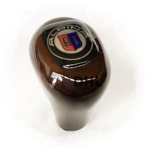 Könnte beinhalten: Ein polierter, dunkelbrauner Holzschaltknauf. Er hat ein rundes Emblem mit dem Wort "ALPINA" und einem Schilddesign in Rot, Blau und Weiß. Der Knauf ist für ein Fahrzeug konzipiert und hat eine glänzende Oberfläche.