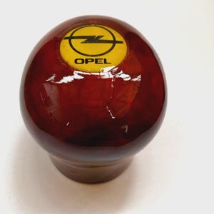 Puede incluir: Pomo de cambio redondo de madera con un acabado brillante de color marrón rojizo. El pomo presenta un logotipo amarillo de Opel con un diseño de rayo negro. La base del pomo es de color marrón claro.