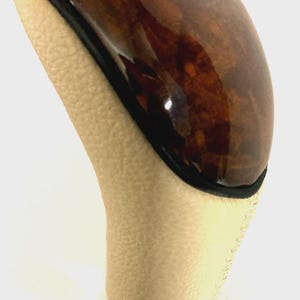 May include: A gear shift knob featuring a polished brown burl wood top with a blue and silver circular emblem. The lower section is covered in textured beige leather, finished with a black base and a thin black trim separating the wood and leather.
