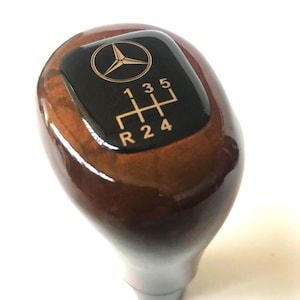 Può includere: Pomello del cambio in legno con finitura lucida. La parte superiore presenta una placca nera con il logo Mercedes-Benz e lo schema del cambio: 1, 3, 5, R, 2 e 4. Il pomello è marrone con un aspetto liscio e lucido.