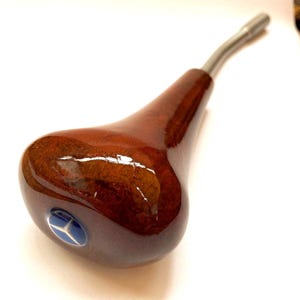 May include: A wooden gear shift knob with a glossy finish. The knob is a dark brown color with a silver metal stem. A blue emblem with a white star is embedded in the base of the knob.