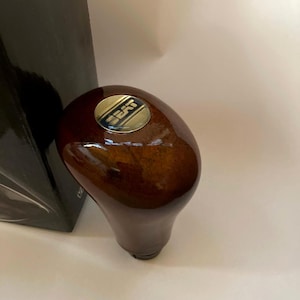 Puede incluir: Un pomo de palanca de cambios de madera con acabado brillante. El pomo es marrón con un emblema metálico que dice "SEAT". El pomo de la palanca de cambios es una pieza de automóvil.