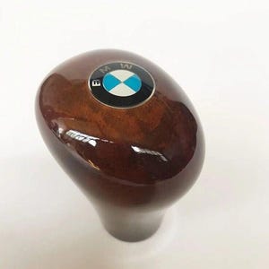 May include: A glossy, brown wooden gear shift knob with a BMW logo on top. The logo features a blue and white circular emblem with the BMW letters around the edge. The knob has a smooth, rounded shape and a dark brown base.