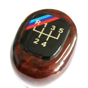 May include: A glossy, wood-grain style shift knob with a black top. The top features a gear shift pattern with numbers and an "R" for reverse. A blue, red, and white stripe design is above the gear pattern.