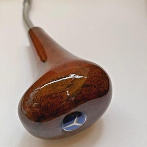Puede incluir: Un pomo de palanca de cambios de madera con acabado brillante. El pomo es de color marrón oscuro con un vástago de metal plateado. Un emblema azul con una estrella blanca es visible en la base del pomo.