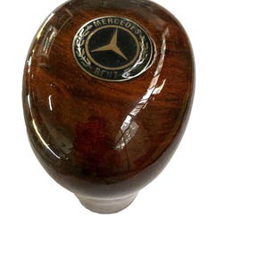 Könnte beinhalten: Ein polierter Schaltknauf aus dunkelbraunem Holzmaserungsmaterial mit einem kreisförmigen Mercedes-Benz-Emblem auf der Oberseite. Das Emblem zeigt einen schwarzen Hintergrund mit einem goldenen Stern, goldener Schrift und einem Lorbeerkranz.