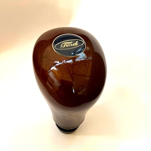 May include: A glossy, dark brown Ford shift knob. The knob has a rounded top with a small, oval emblem featuring the Ford logo in white text on a blue background. The base of the knob is black.
