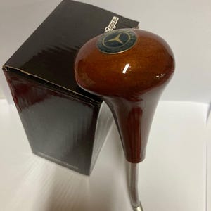 May include: A brown wooden gear shift knob with a glossy finish, featuring a Mercedes-Benz emblem. The knob has a silver metal shaft and is next to a black box.