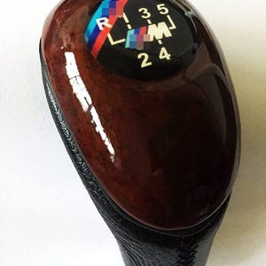 May include: A car gear shift knob with a dark wood grain finish and a black leather base. The top features a black dial with white numbers and the BMW logo in blue, red, and white.