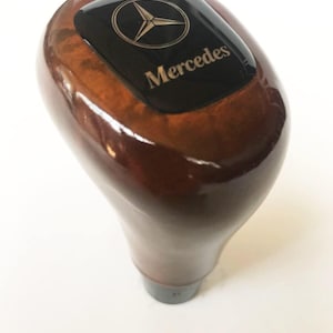 May include: A polished, brown wooden gear stick knob with a black rectangular top. The top features the Mercedes-Benz logo and brand name in gold lettering. The knob has a smooth, ergonomic design.