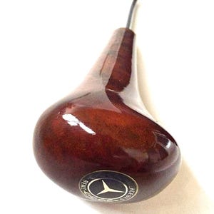 May include: A dark brown, pear-shaped gear shift knob with a shiny finish. The knob has a metal stem and a circular emblem with the Mercedes-Benz logo. The emblem is silver and black.