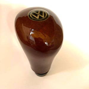 May include: A brown, wooden gear shift knob with a glossy finish. The top of the knob features a gold and black Volkswagen logo. The knob has a rounded top and a tapered base.