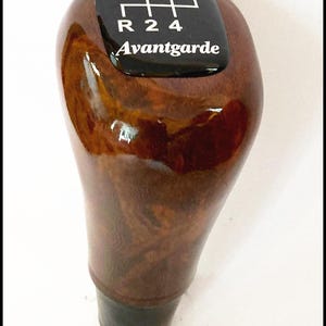 Op de afbeelding: Een houten versnellingspookknop met een glanzende afwerking. De bovenkant heeft een zwart paneel met een wit schakelpatroon en het woord "Avantgarde". De knop heeft een donkerbruin houtnerfpatroon.