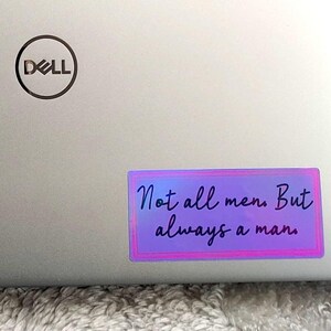 Könnte beinhalten: Ein holografischer Aufkleber mit einem lila und rosa Rand, der auf einem silbernen Laptop angebracht ist. Der Aufkleber lautet "Not all men. But always a man" in einer handschriftlichen Schriftart.