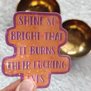 Könnte beinhalten: Ein orangefarbener Aufkleber mit dem Text "SHINE SO BRIGHT THAT IT BURNS THEIR FUCKING EYES. YES." Der Aufkleber hat einen lila Rand und Sternakzente. Im Hintergrund sind zwei goldene Schalen.