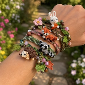 May include: A multi-layered braided bracelet featuring small crocheted animal figures including a white rabbit with a carrot, an orange fox, a grey raccoon, and a brown hedgehog, accented with green crocheted leaves, pink beads, and small charms.