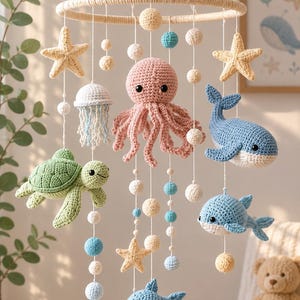 Op de afbeelding: Een hangende mobiel met gehaakte zeedieren, waaronder een roze octopus, een groene schildpad, twee blauwe walvissen, een witte kwal en twee gele zeesterren. De figuren hangen aan een rond frame met kleine witte en blauwe wollen bolletjes.