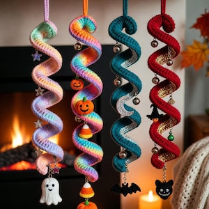 May include: Four colorful, crochet spiral ornaments hang from a wooden mantel. Each has a different color scheme and Halloween-themed charms, including ghosts, pumpkins, bats, and bells. The background features a fireplace, candles, and fall leaves.