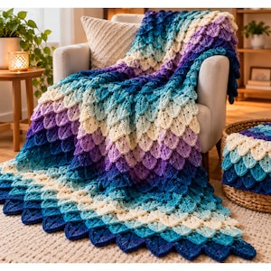 May include: A crochet blanket featuring a textured scale pattern in a gradient of colors, transitioning from deep navy blue and royal blue to light turquoise, pale blue, cream, and shades of purple, draped over a light-colored armchair.