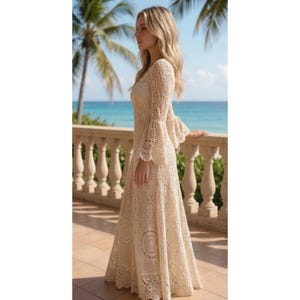 May include: A cream-colored, long crochet dress with bell sleeves. The dress features a V-neck and a flowing skirt with circular patterns. The dress is made of a light, airy material, perfect for a beach or outdoor event. The background shows a tropical scene with palm trees and ocean.