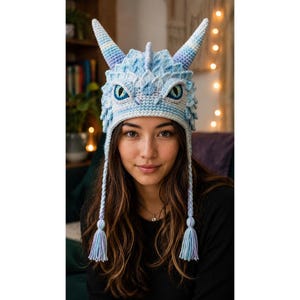 Peut inclure: Un bonnet dragon crocheté bleu clair et blanc avec deux cornes proéminentes, un motif d'écailles texturé et deux longues oreillettes tressées terminées par des pompons. Le bonnet présente des yeux détaillés avec des pupilles sombres.