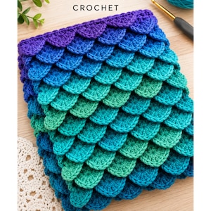 Puede incluir: Una pieza de ganchillo con un patrón de escamas texturizado en un degradado de colores que va desde el morado oscuro en la parte superior hasta el azul, turquesa y verde. La palabra CROCHET está impresa en negro arriba y se ve un ganchillo.