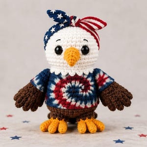 May include: A crocheted eagle doll with a patriotic theme. The eagle has a white head, a yellow beak, and large black eyes. It wears a blue and red tie-dye shirt and a headband with stars and stripes. The eagle's wings and feet are brown and yellow.