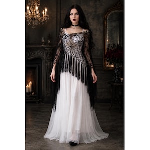 May include: A floor-length white dress featuring a sleeveless bodice and a flowing skirt, paired with a black, sheer, spiderweb-patterned shawl that has long, dangling fringe trim along the bottom edge and a black choker necklace.
