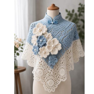 May include: A light blue and cream crochet shawl featuring a high mandarin collar with a single button closure. The garment is adorned with layered, three-dimensional crochet flowers in matching blue and cream tones, each centred with a white pearl bead.
