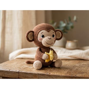 May include: A crocheted monkey doll in shades of brown and beige, holding a yellow banana. The handmade toy has a friendly expression with rosy cheeks and large eyes. It is sitting on a wooden surface.