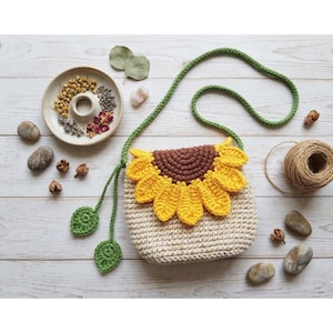 Puede incluir: Bolso bandolera de ganchillo con diseño de girasol. El bolso tiene un cuerpo color crema, un centro marrón y pétalos amarillos. Tiene una correa verde y dos detalles de hojas verdes. El bolso está hecho a mano y está sobre una superficie de madera blanca.