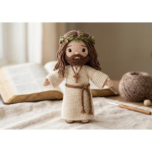 May include: A handmade crochet doll of Jesus, wearing a cream-colored robe, a brown belt, and a wooden cross necklace. The doll has brown hair, a beard, and a crown of green leaves. The doll is approximately 7 inches tall.